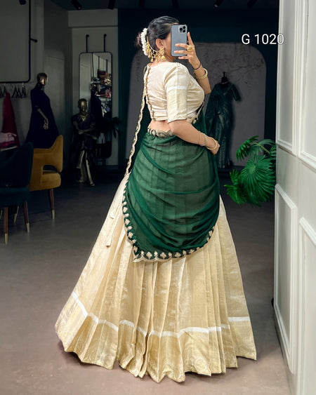 A woman wearing a traditional Indian lehenga choli dress, photographed in a mirror selfie inside what appears to be a boutique or showroom. The outfit features a cream-colored flared skirt (lehenga) with white border details and a dark green draped blouse and dupatta (sari) with a decorative border of small white flowers.