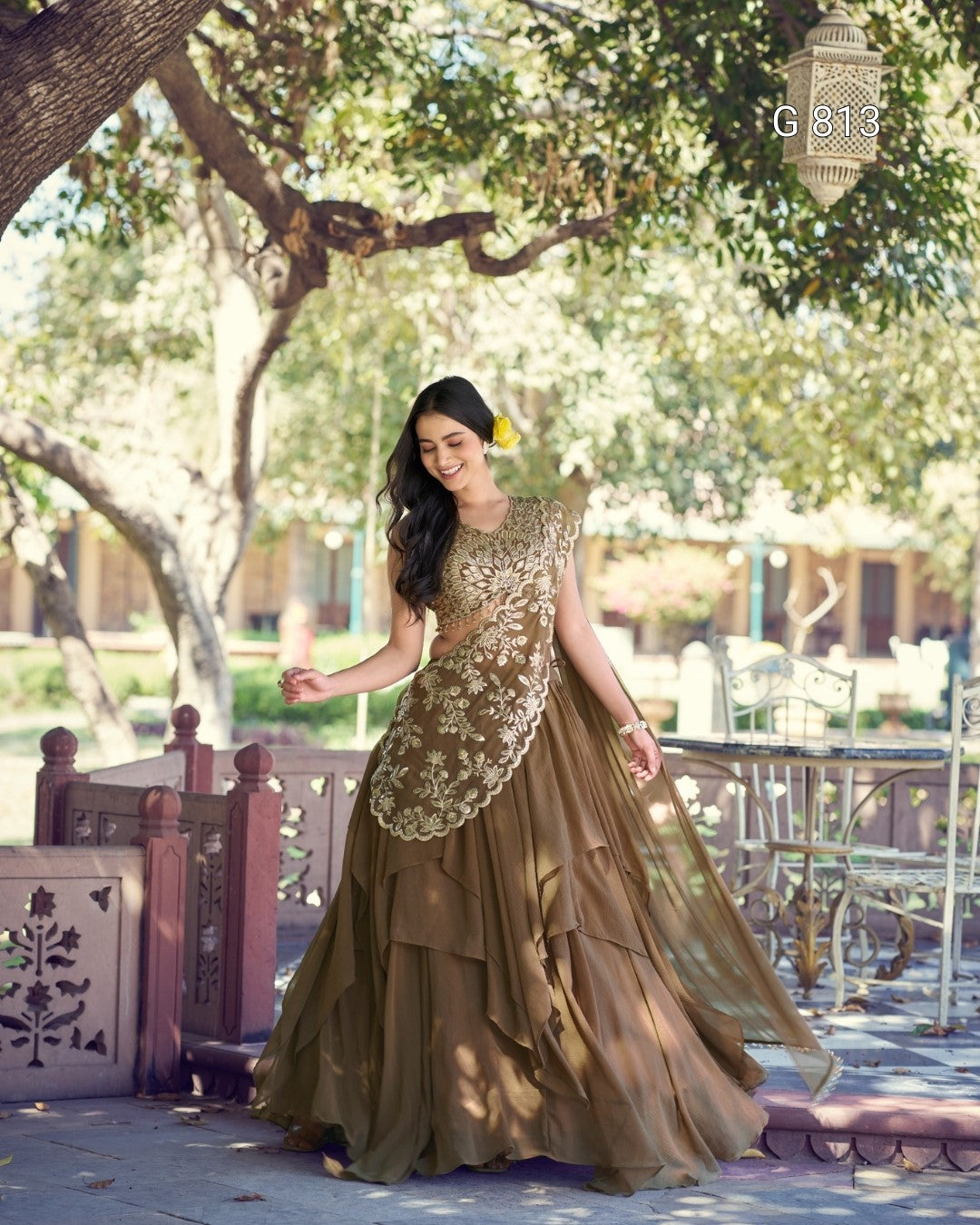 Woman in a brown and gold dress standing outdoors with trees and chairs in the background