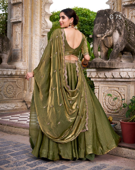 A woman wearing an olive green traditional Indian lehenga choli dress with gold embroidery, standing outdoors in front of a stone elephant statue and carved pillars.