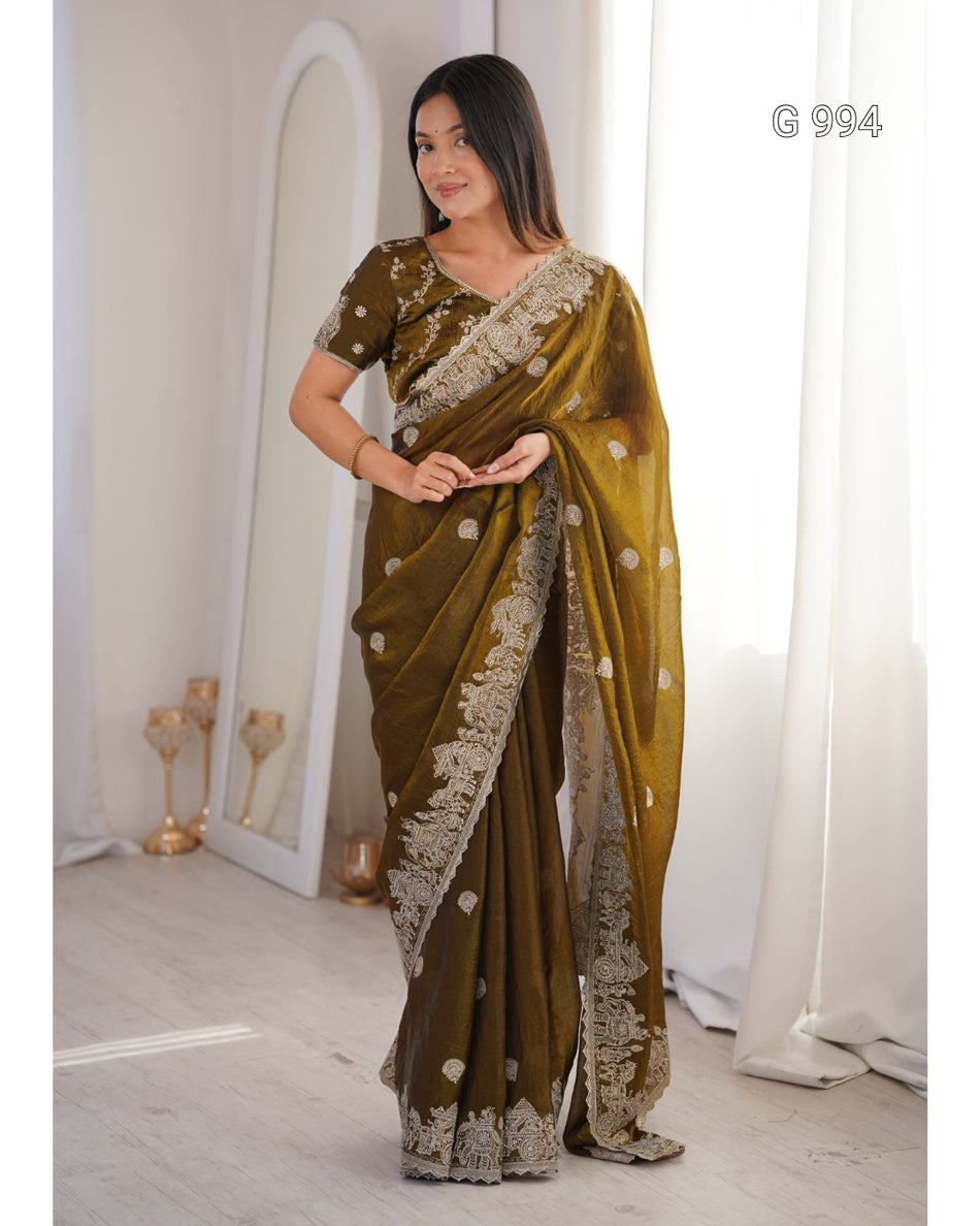 A woman modeling an olive green saree with beige floral embroidery and a matching embroidered blouse, standing in a bright indoor room with white curtains and a mirror.