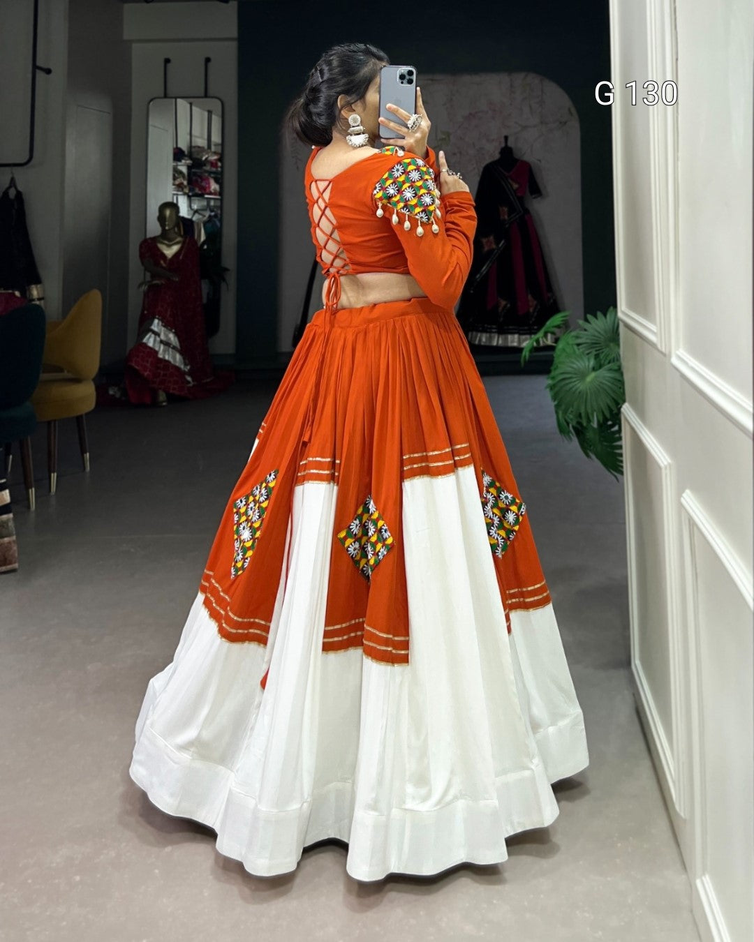 Person wearing an orange and white traditional outfit with intricate designs, standing in a room.