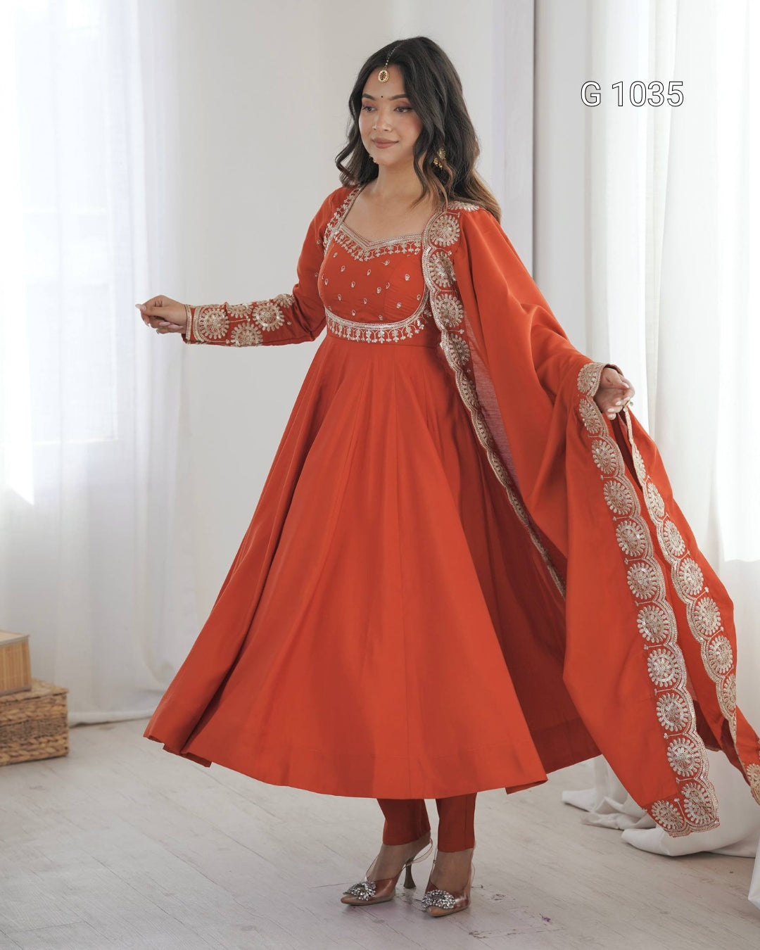 A woman models a vibrant orange-red Anarkali gown with gold embroidery, paired with matching trousers and a draped dupatta, in a bright, minimalist room with white curtains.