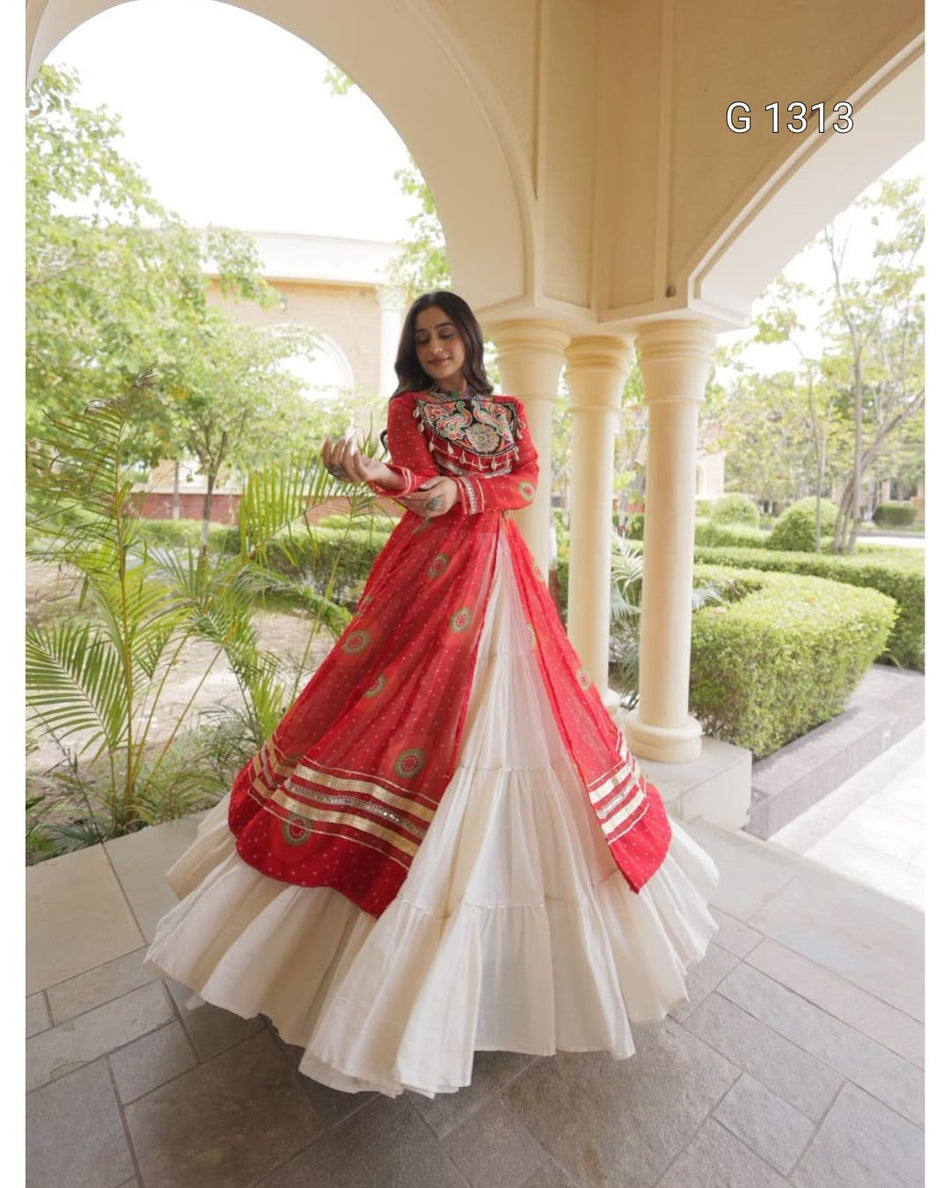 A woman wearing a traditional Indian red and white lehenga choli dress with gold embroidery, standing in a covered outdoor walkway with columns and green garden plants in the background.