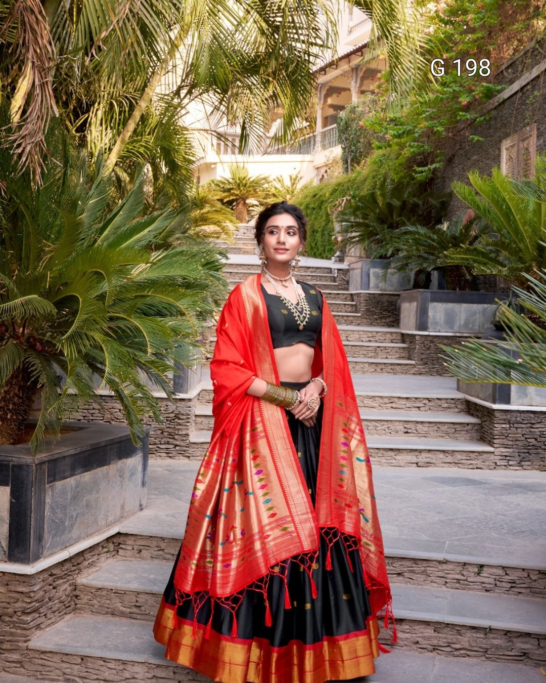 Woman in a red and black traditional outfit standing outdoors with greenery.