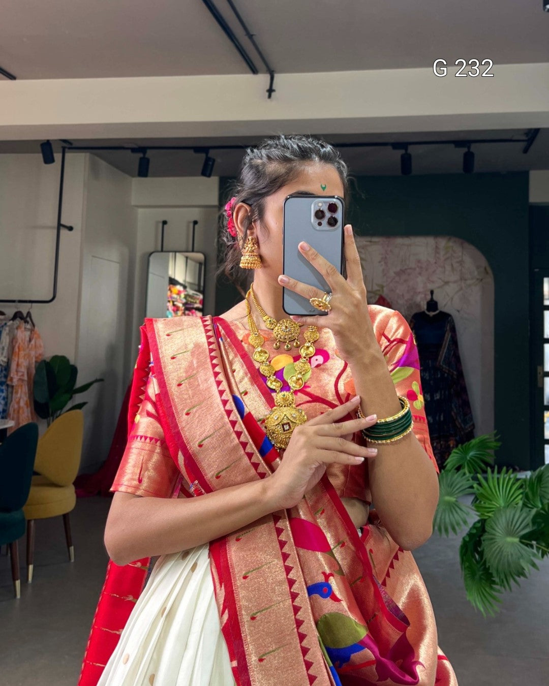 Person wearing a colorful saree with gold jewelry, taking a mirror selfie.