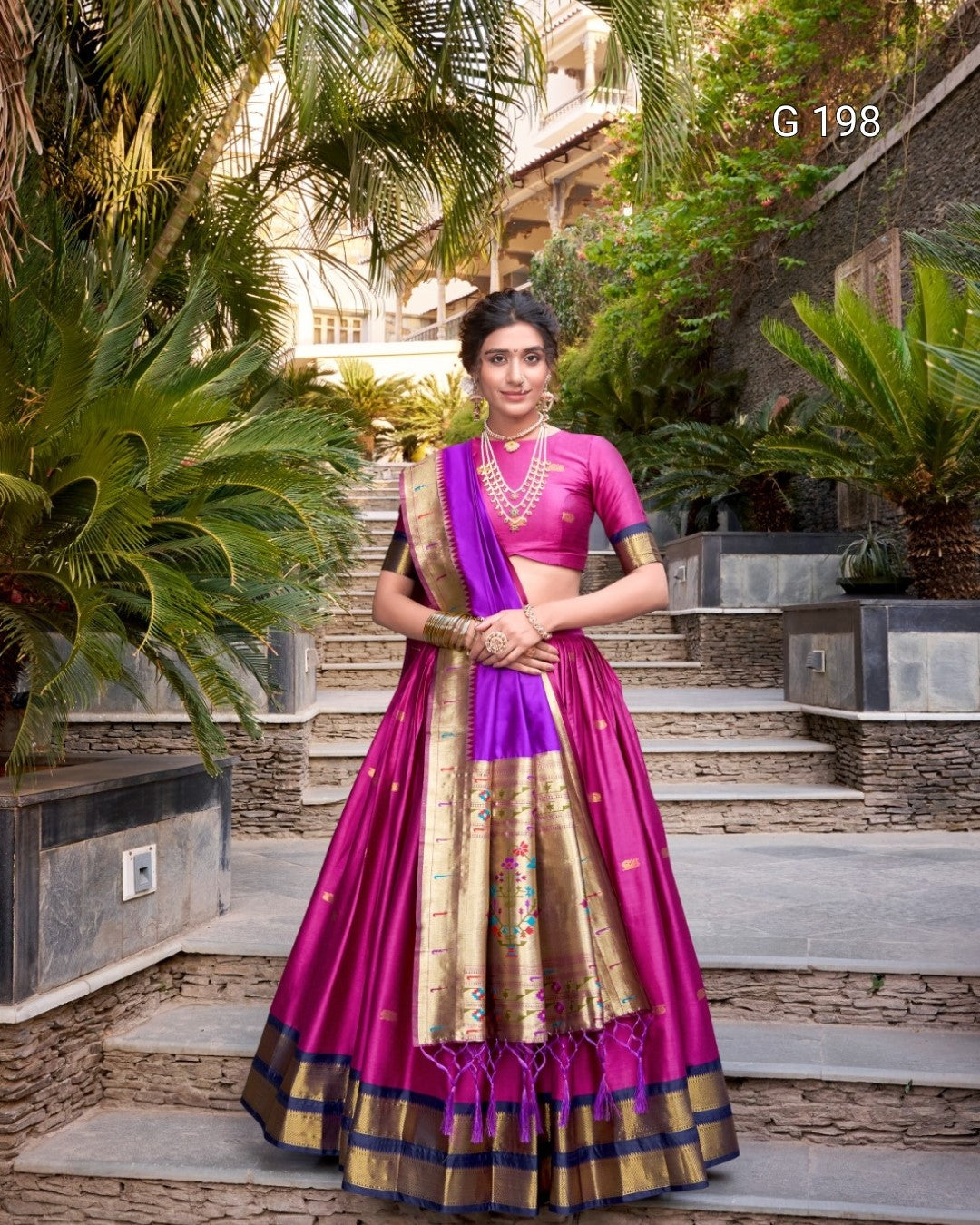 Woman in a traditional purple and gold saree standing outdoors with greenery.