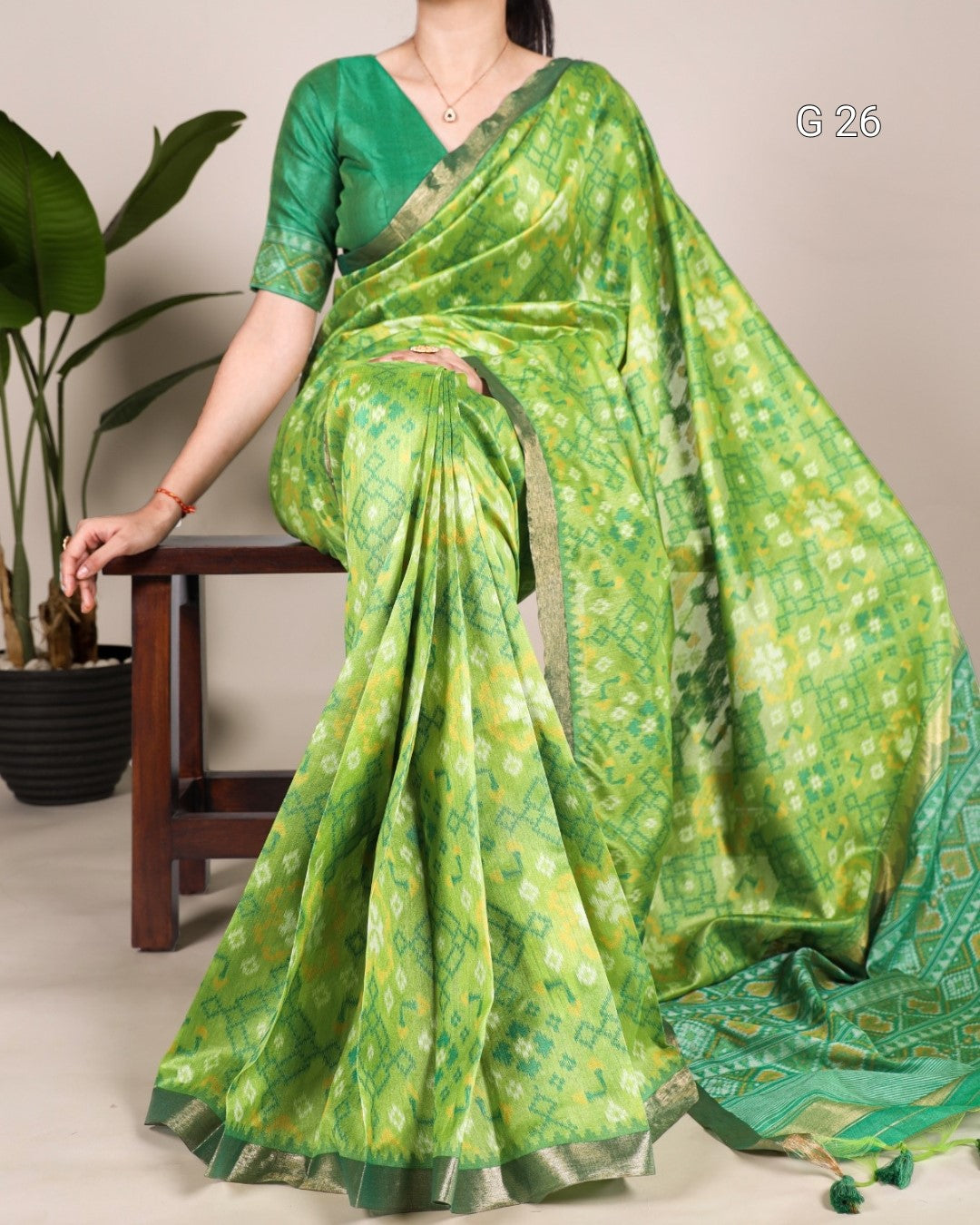 Green saree with a pattern draped over a person, sitting on a chair with a plant in the background.