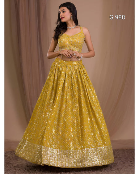 A woman modeling a yellow embellished lehenga choli set against a beige draped backdrop. The set features a sleeveless yellow top with intricate gold embroidery and a matching full skirt with a wide gold zari border.