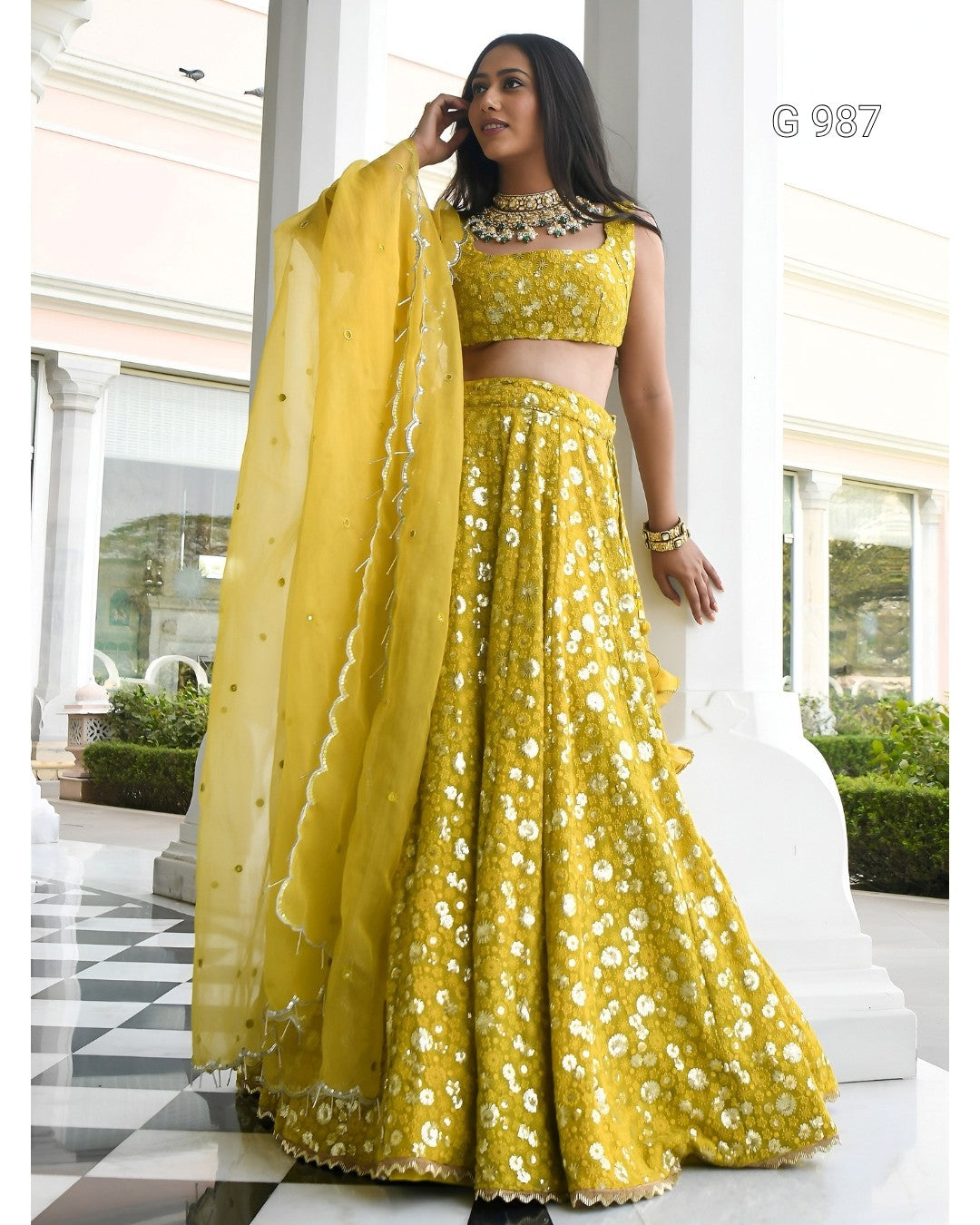 A woman models a bright yellow Indian ethnic wear set consisting of a sleeveless crop top, flared skirt, and matching dupatta, decorated with white floral embroidery, in an outdoor colonnade setting.
