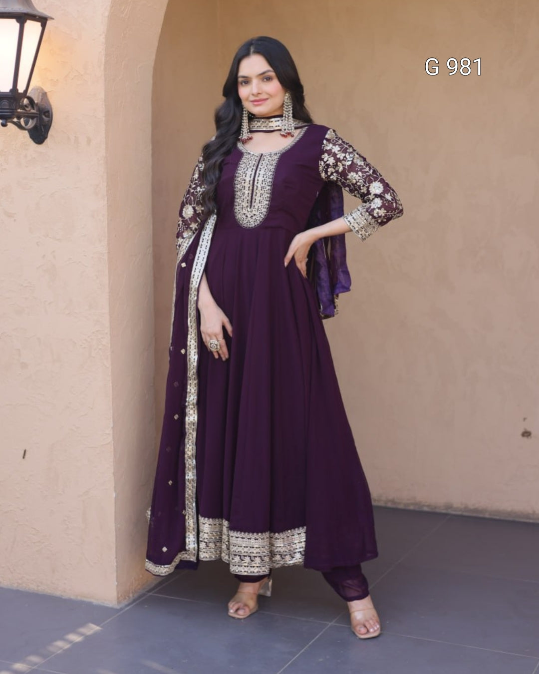 A woman models a deep purple Anarkali gown with silver and gold embroidery, paired with a matching sheer dupatta, set against a beige stucco wall background.