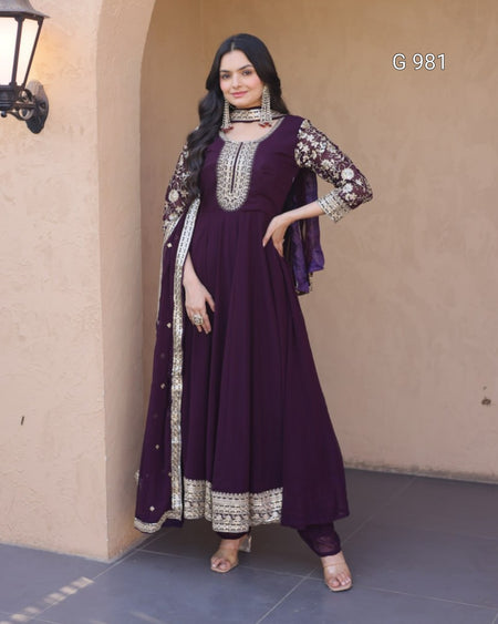 A woman models a deep purple Anarkali gown with silver and gold embroidery, paired with a matching sheer dupatta, set against a beige stucco wall background.