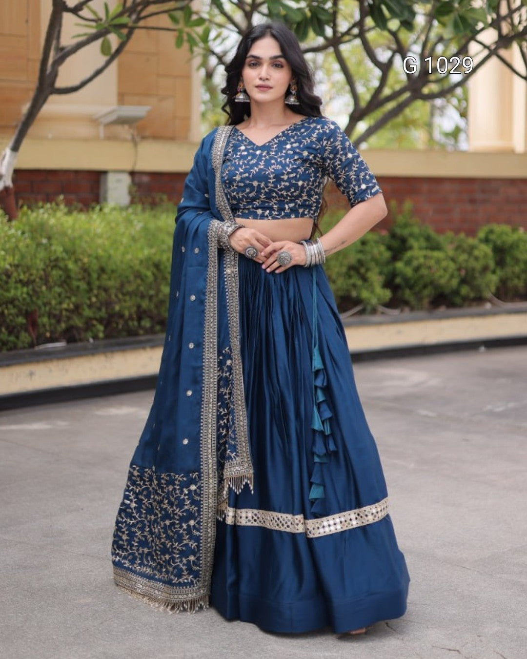 A woman stands outdoors wearing a navy blue traditional outfit with gold embroidery, featuring a flared skirt, embroidered blouse, and matching draped scarf.
