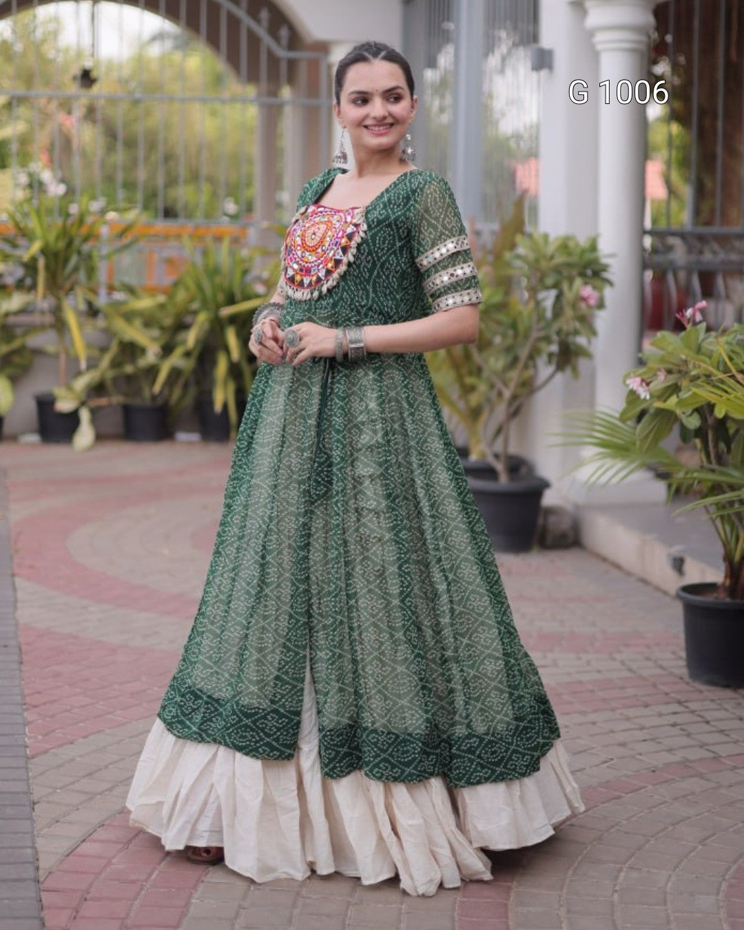 A woman wearing a dark green embroidered Anarkali gown with a cream-colored ruffled underskirt, standing in an outdoor courtyard setting.