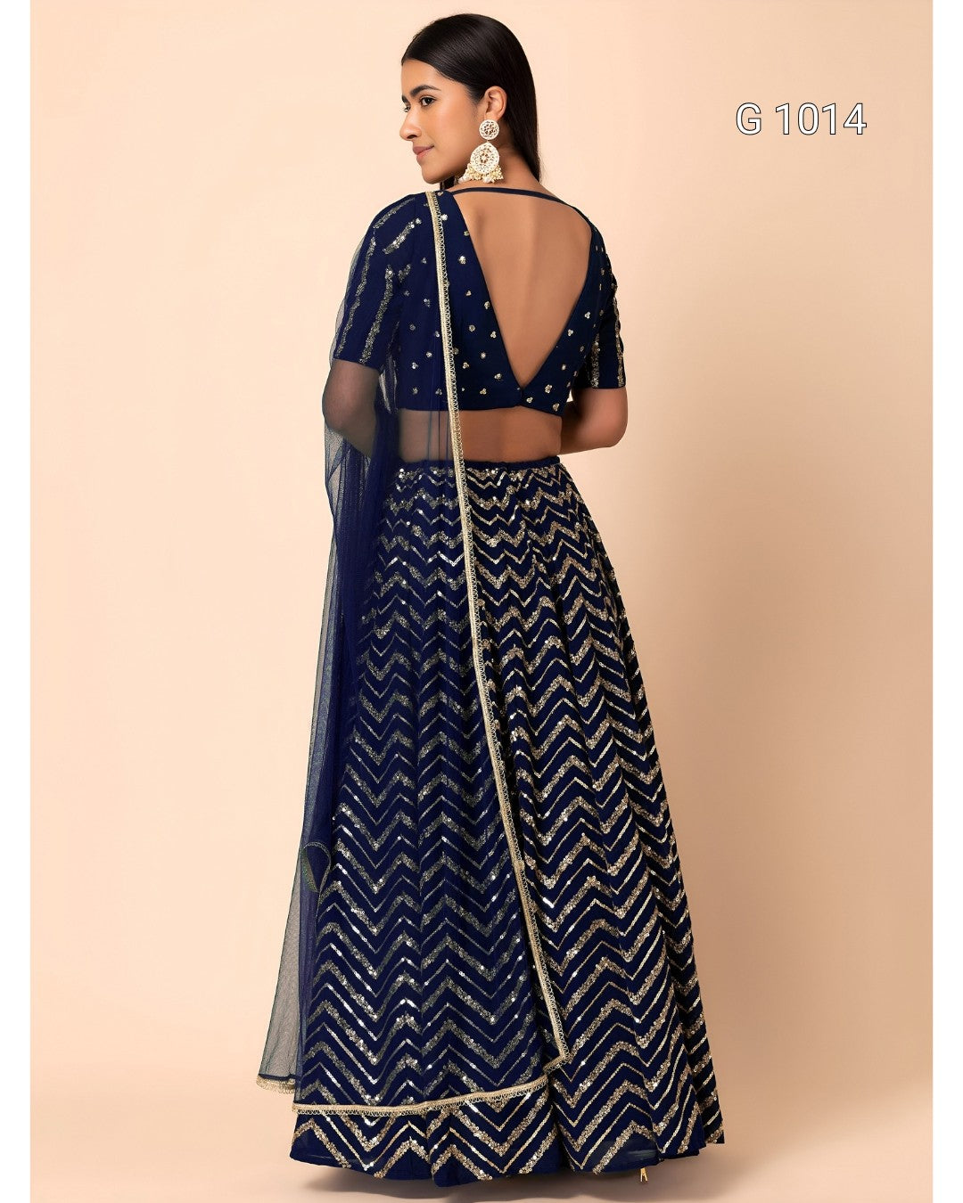 A woman modeling a navy blue and silver embellished lehenga choli set from the back, featuring a deep open back, silver zari chevron patterns, and a sheer dupatta, set against a solid beige background.