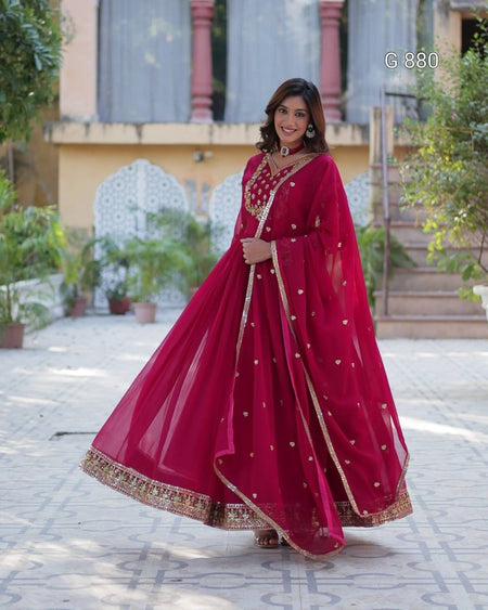 Woman in a red traditional outfit standing outdoors with plants and a building in the background.