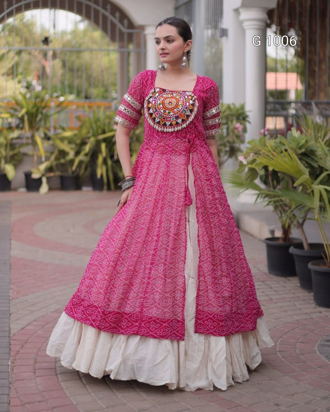 A woman wearing a bright pink embroidered Anarkali gown with a white ruffled underskirt, standing in an outdoor courtyard with potted plants.