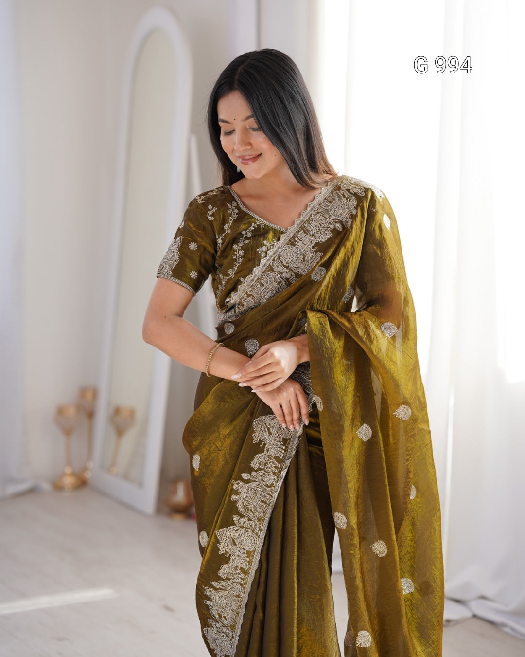 A woman wearing an olive green saree with intricate silver embroidery and a matching embroidered blouse, standing in a bright, minimalist room with white curtains and a mirror.