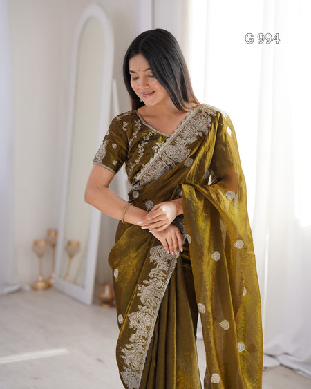 A woman wearing an olive green saree with intricate silver embroidery and a matching embroidered blouse, standing in a bright, minimalist room with white curtains and a mirror.