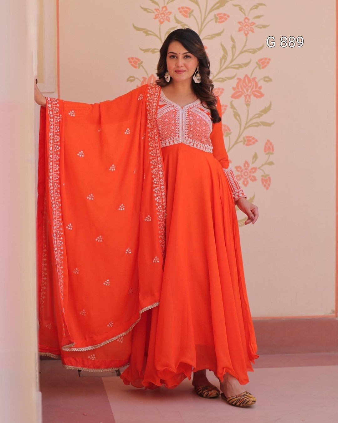 Woman wearing an orange traditional outfit with white embroidery against a floral wall.