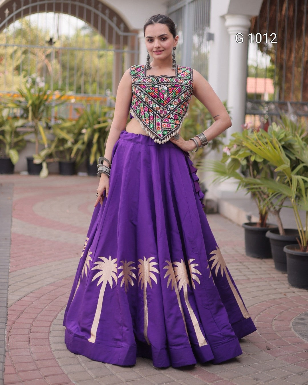 A woman modeling a traditional Indian ethnic wear set featuring a purple flared skirt with gold palm tree embroidery and a matching embroidered sleeveless blouse, standing in an outdoor courtyard.