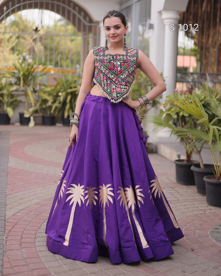 A woman modeling a traditional Indian ethnic wear set featuring a purple flared skirt with gold palm tree embroidery and a matching embroidered sleeveless blouse, standing in an outdoor courtyard.