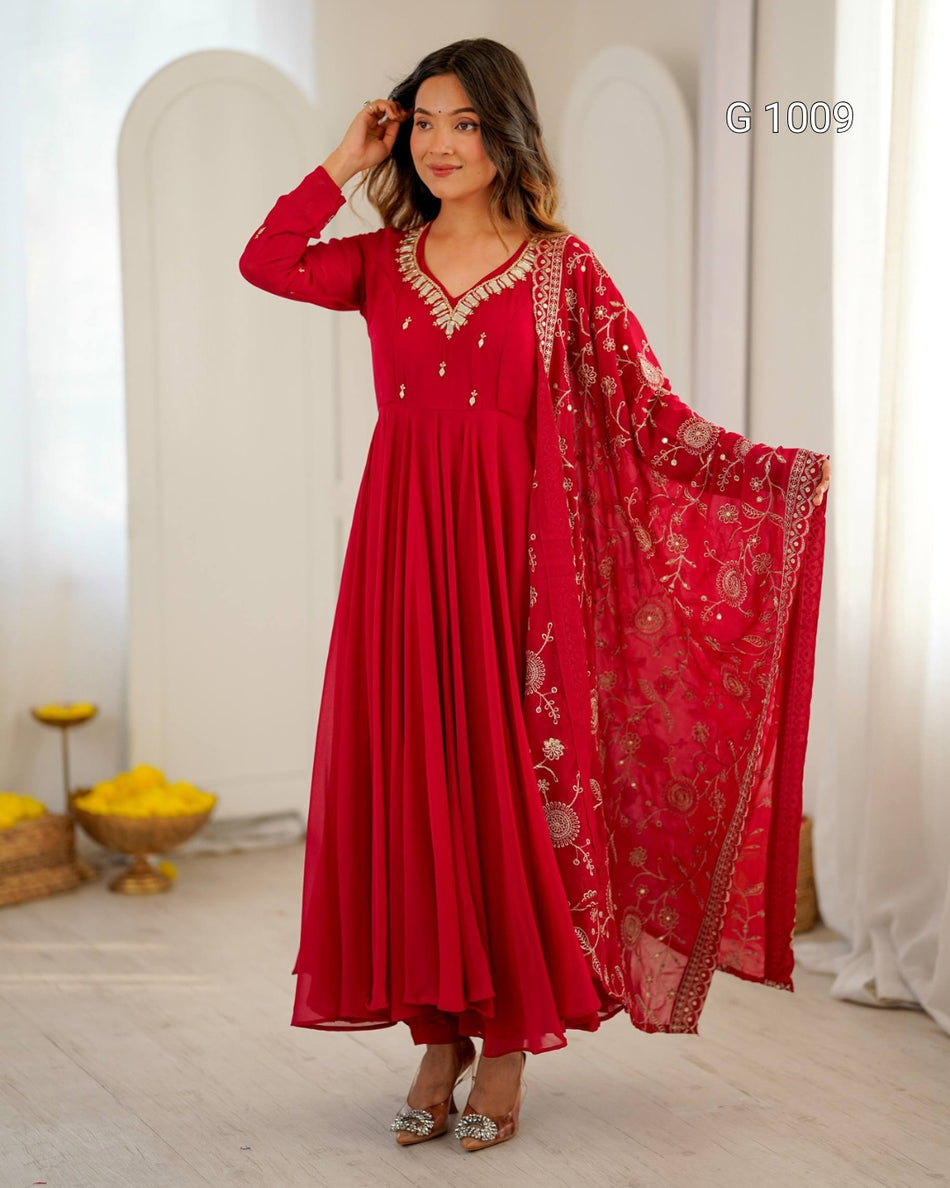 A woman modeling a long red dress with gold embroidery and a matching sheer dupatta draped over her shoulder, set against a bright, minimalist indoor background with white arches.