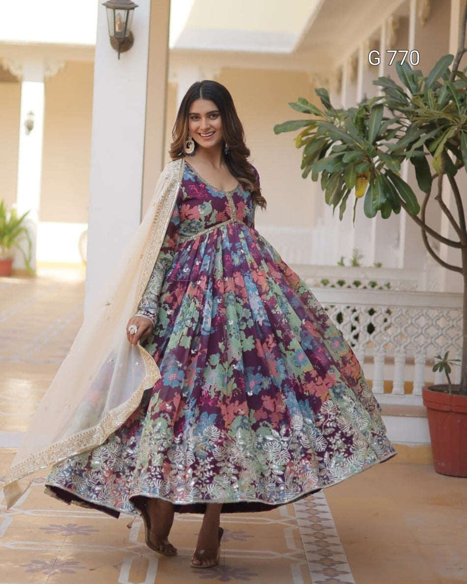 Woman in a colorful floral dress standing in an outdoor setting with plants and architectural elements.