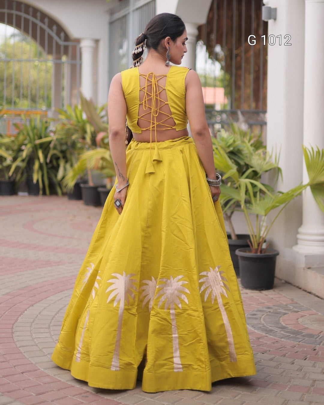 A woman wearing a bright yellow traditional dress with a flared skirt and lace-up back, photographed from behind in an outdoor courtyard setting with potted plants.