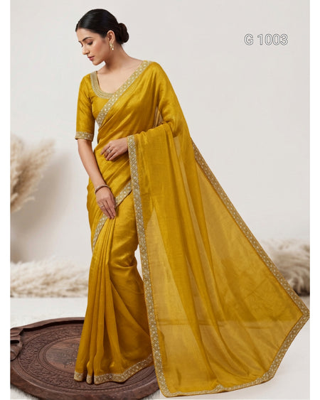 A woman models a mustard yellow saree with gold embroidered borders and a matching embroidered blouse, standing on a round wooden platform against a light beige background.