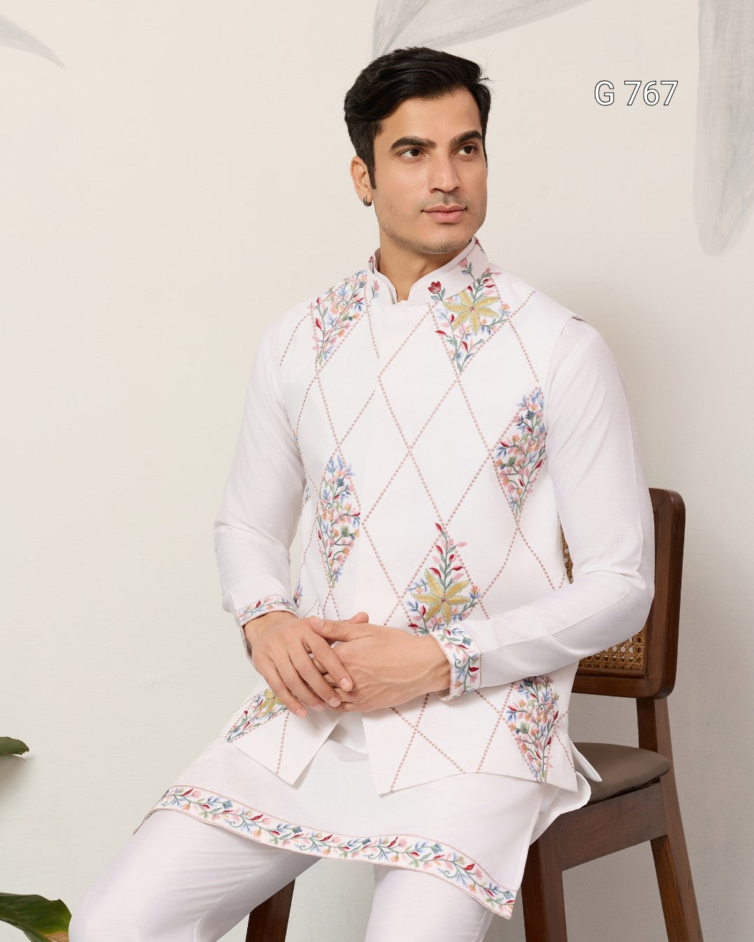 Man wearing a white embroidered traditional outfit sitting on a chair against a plain background