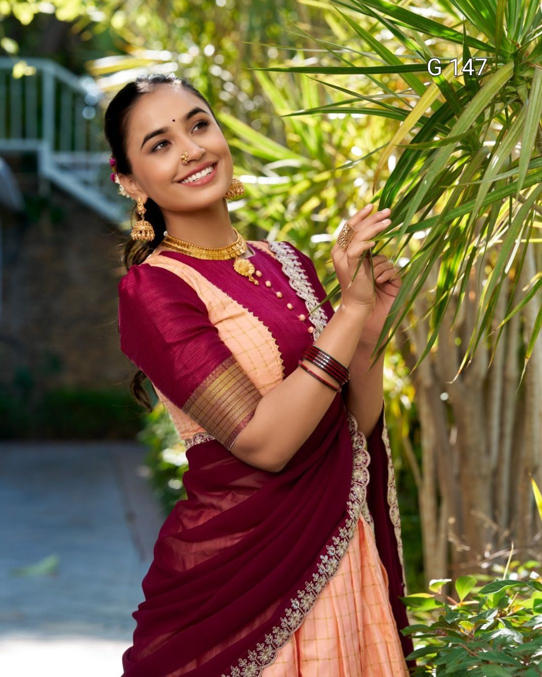 Woman in a traditional maroon and peach saree standing among green plants.