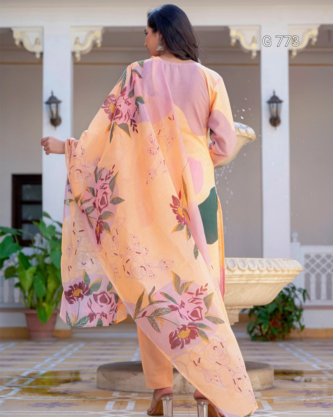 Woman wearing a floral outfit in an outdoor setting with a fountain and plants.