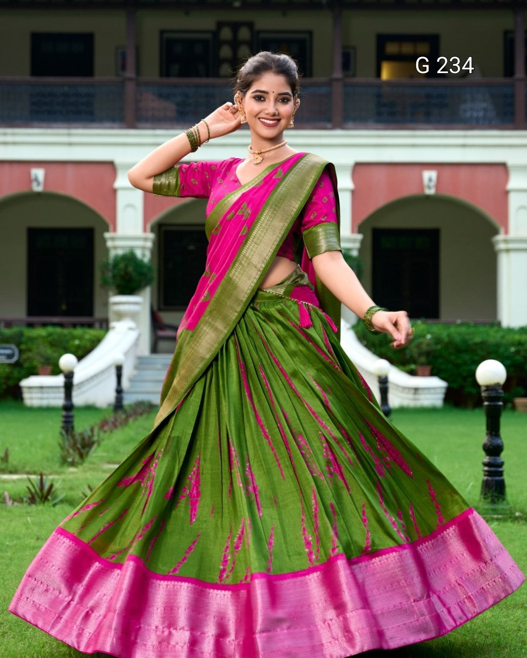 Woman in a green and pink saree standing outdoors with a building in the background