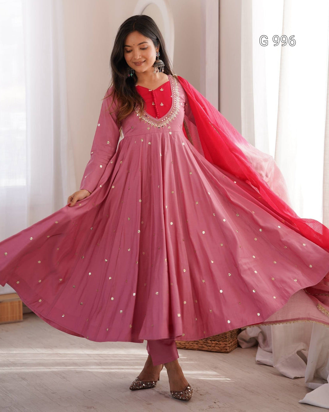 A woman modeling a pink traditional Anarkali gown with a red embroidered neckline and a bright red dupatta, standing in a softly lit indoor setting.