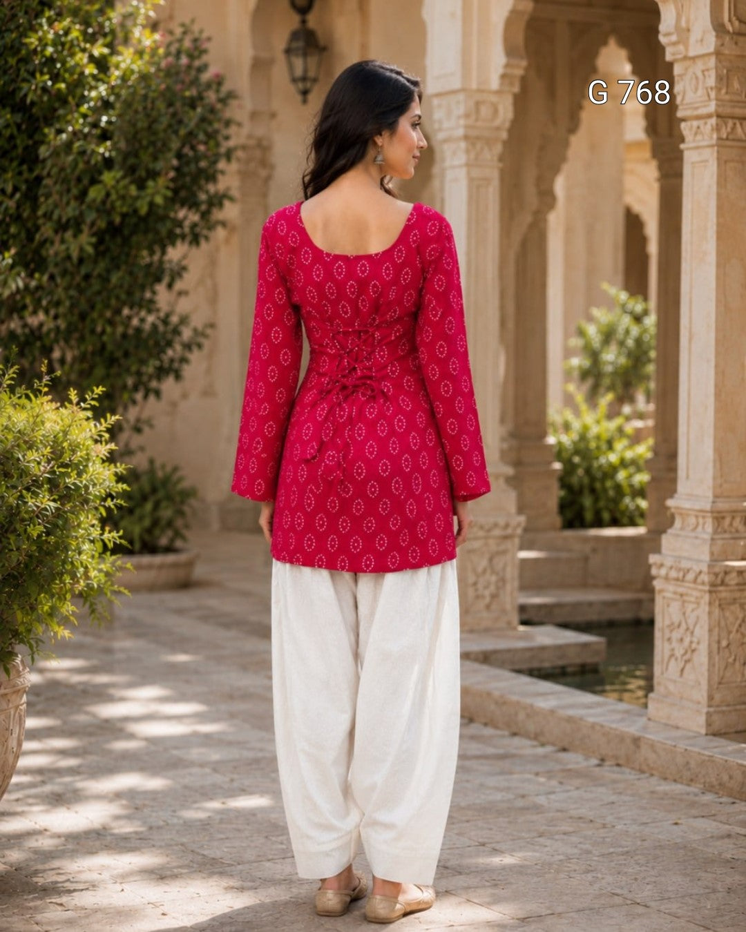 Woman wearing a red patterned top and white pants standing in an outdoor setting with architectural elements.