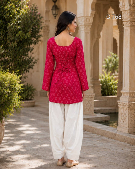 Woman wearing a red patterned top and white pants standing in an outdoor setting with architectural elements.