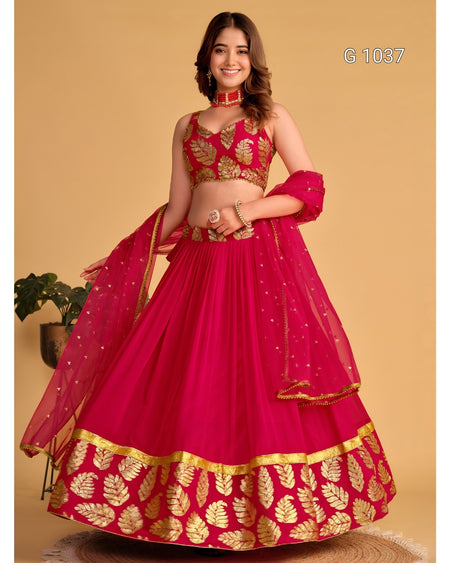 A woman models a bright pink lehenga choli set with gold leaf embroidery, featuring a flared skirt, embroidered blouse, and matching dupatta against a solid beige background.