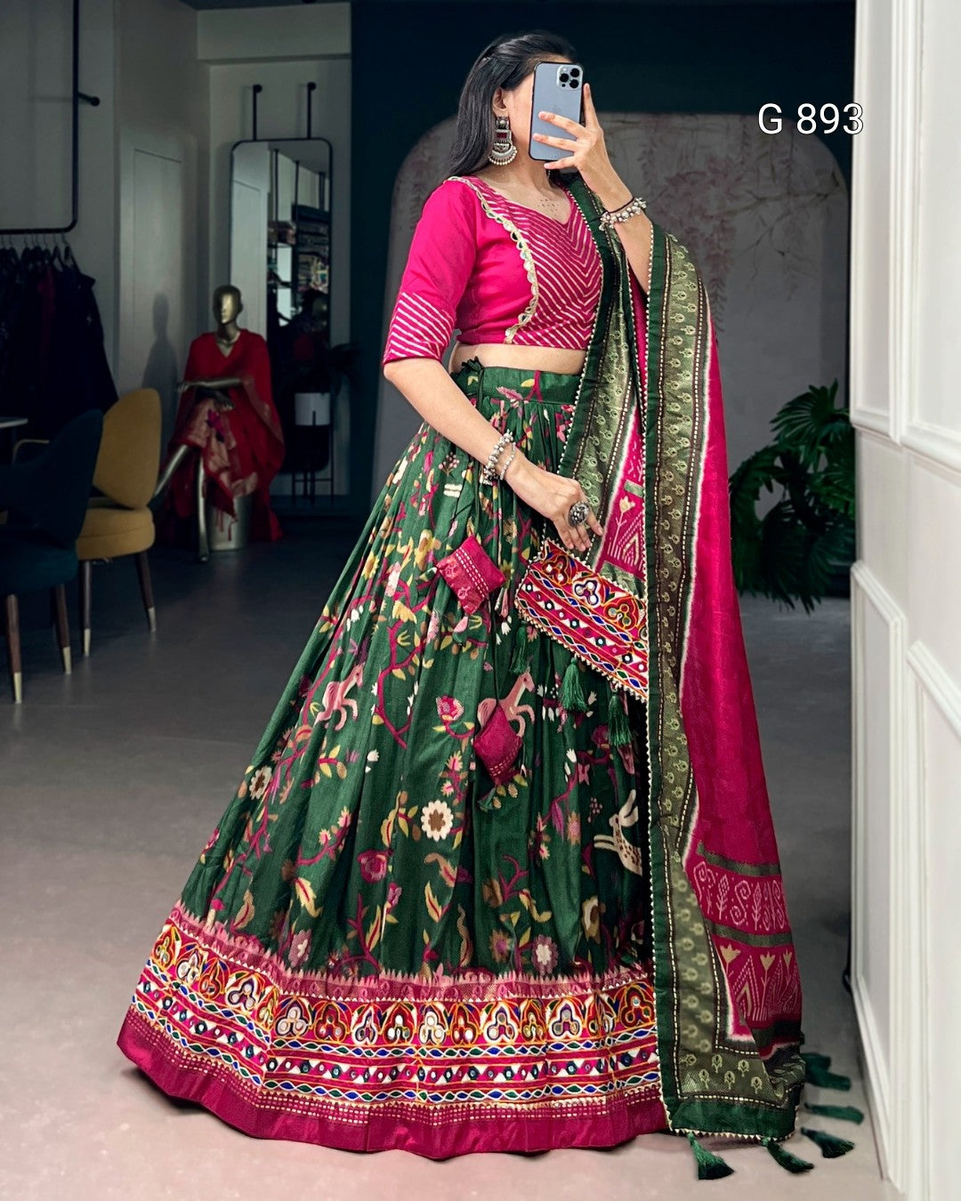 Woman wearing a green and pink traditional outfit with a patterned dupatta, taking a mirror selfie.