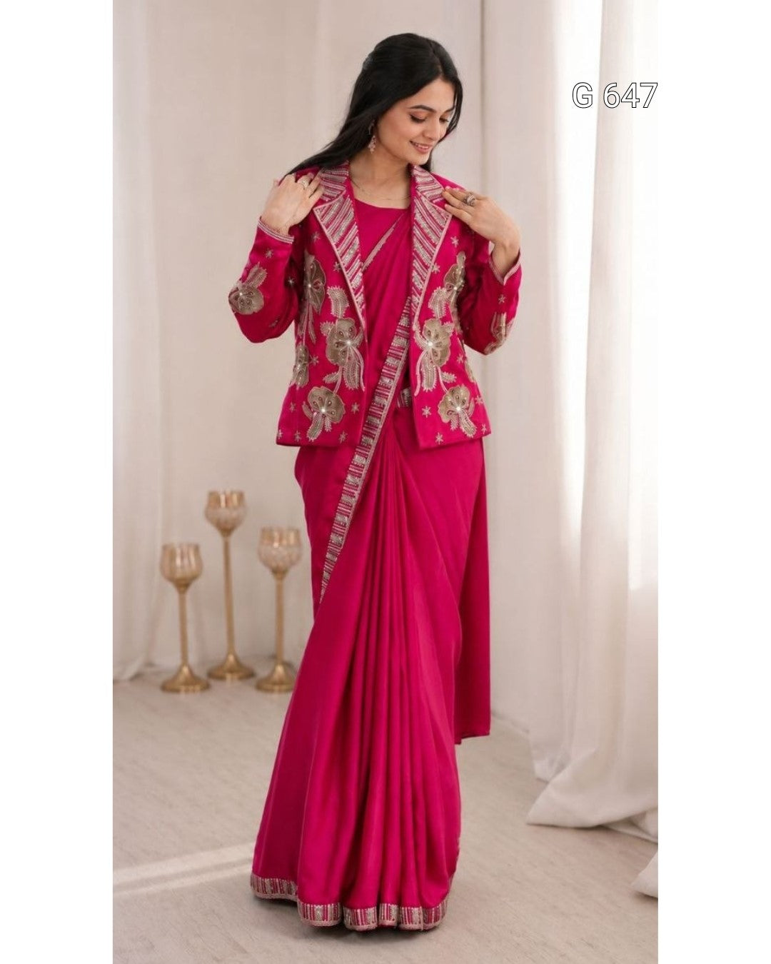 A woman modeling a bright pink traditional outfit with a matching draped skirt, decorated with ornate gold and silver floral embroidery and patterned borders. She is standing in a softly lit indoor setting with white curtains and decorative gold candle holders.