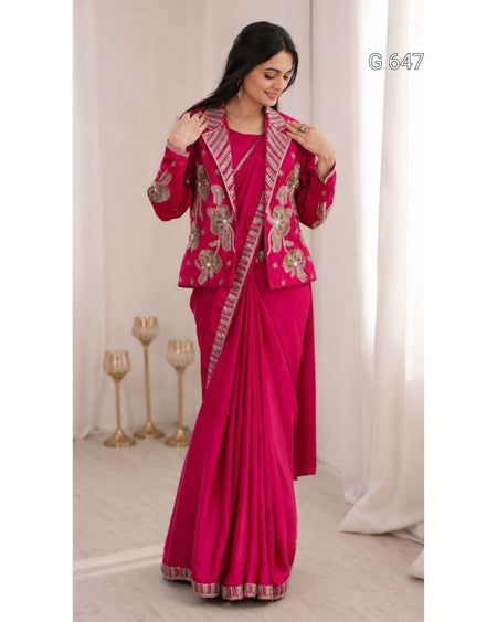 A woman modeling a bright pink traditional outfit with a matching draped skirt, decorated with ornate gold and silver floral embroidery and patterned borders. She is standing in a softly lit indoor setting with white curtains and decorative gold candle holders.