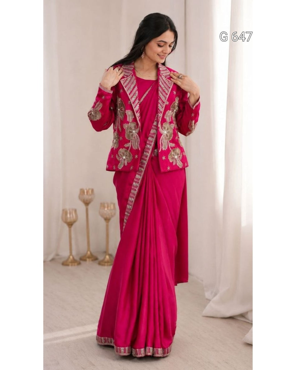 A woman modeling a bright pink traditional outfit with a matching draped skirt, decorated with ornate gold and silver floral embroidery and patterned borders. She is standing in a softly lit indoor setting with white curtains and decorative gold candle holders.