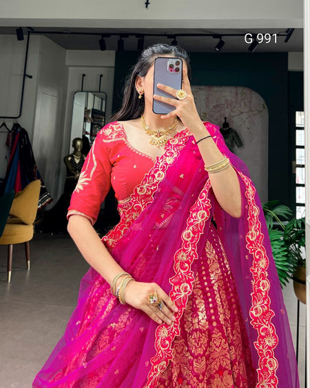 A woman takes a mirror selfie wearing a bright pink lehenga choli with intricate gold embroidery. The setting appears to be a mirror selfie in an indoor boutique or fitting space, with clothing racks, mirrors, and a plant visible in the background.