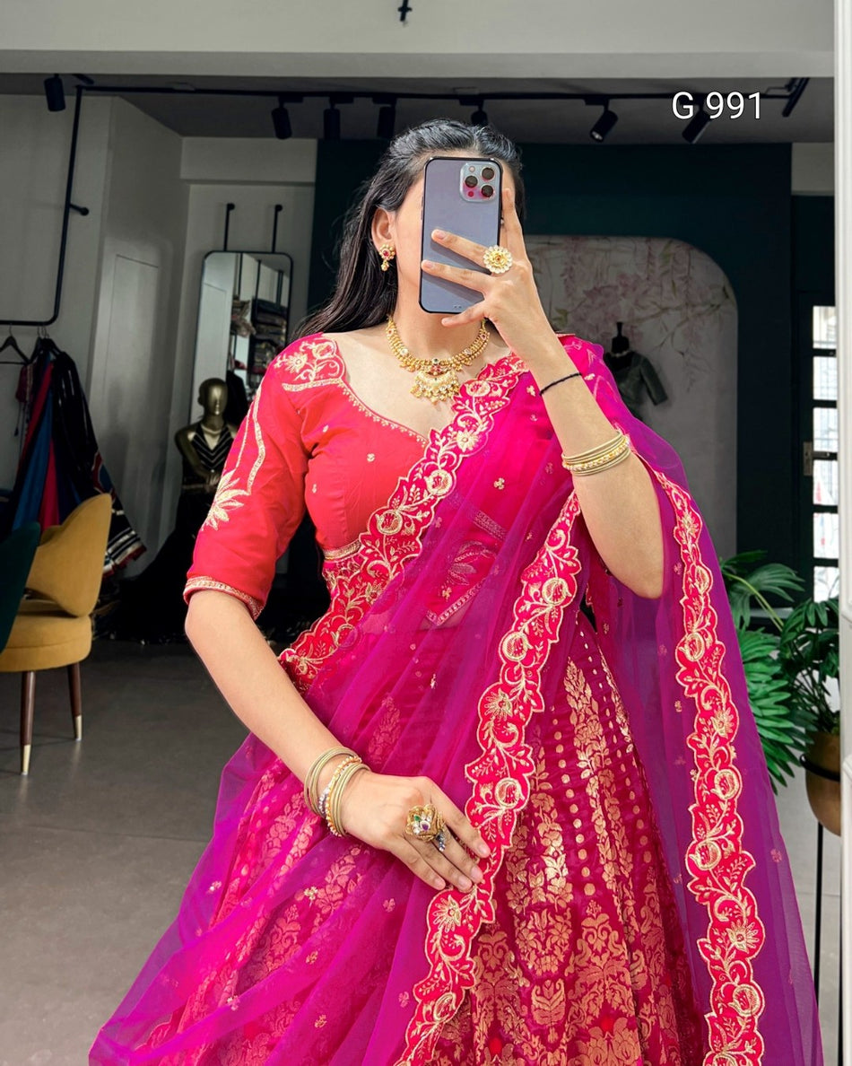 A woman takes a mirror selfie wearing a bright pink lehenga choli with intricate gold embroidery. The setting appears to be a mirror selfie in an indoor boutique or fitting space, with clothing racks, mirrors, and a plant visible in the background.