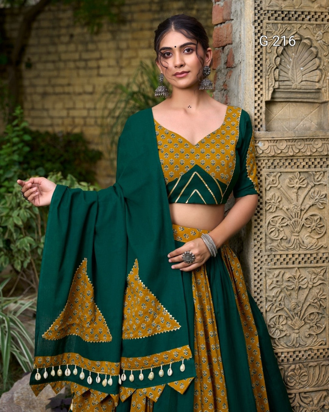 Woman wearing a green and yellow traditional outfit with a patterned dupatta, standing against an architectural background.