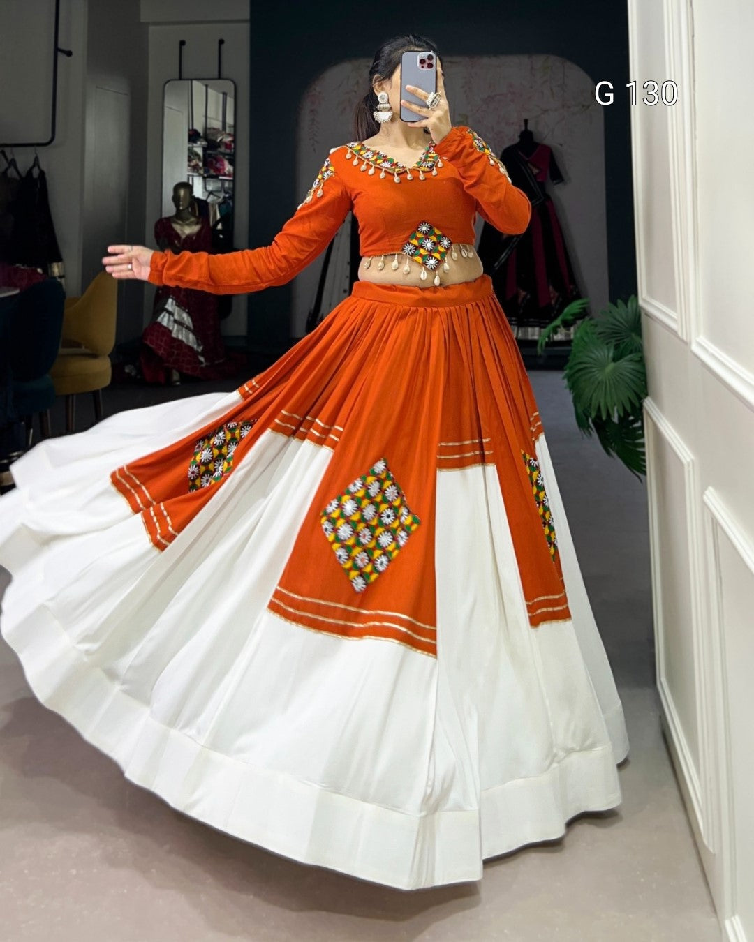 Person wearing an orange and white traditional outfit with intricate designs, standing in a room.