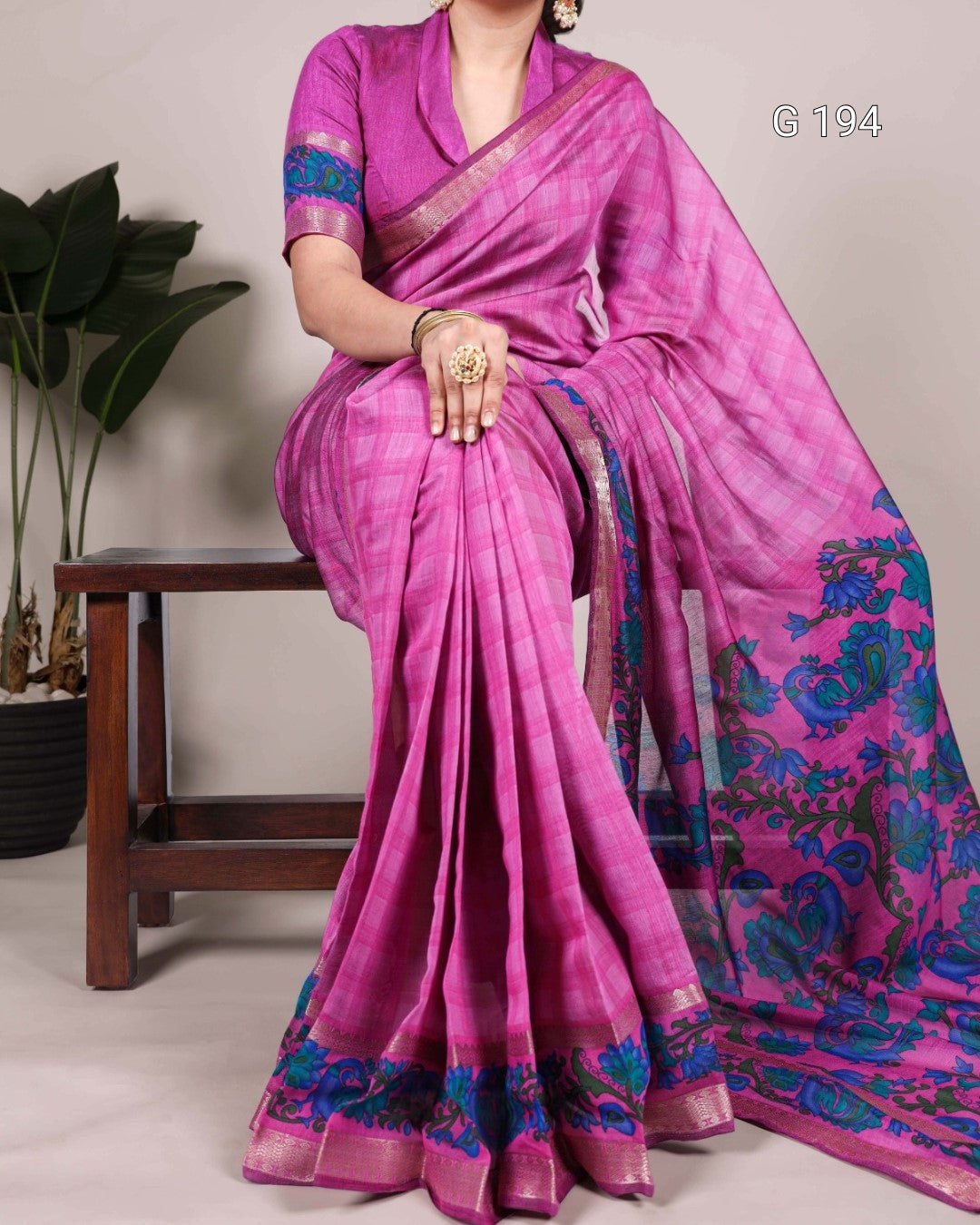 Woman wearing a pink saree with blue patterns, sitting on a chair.