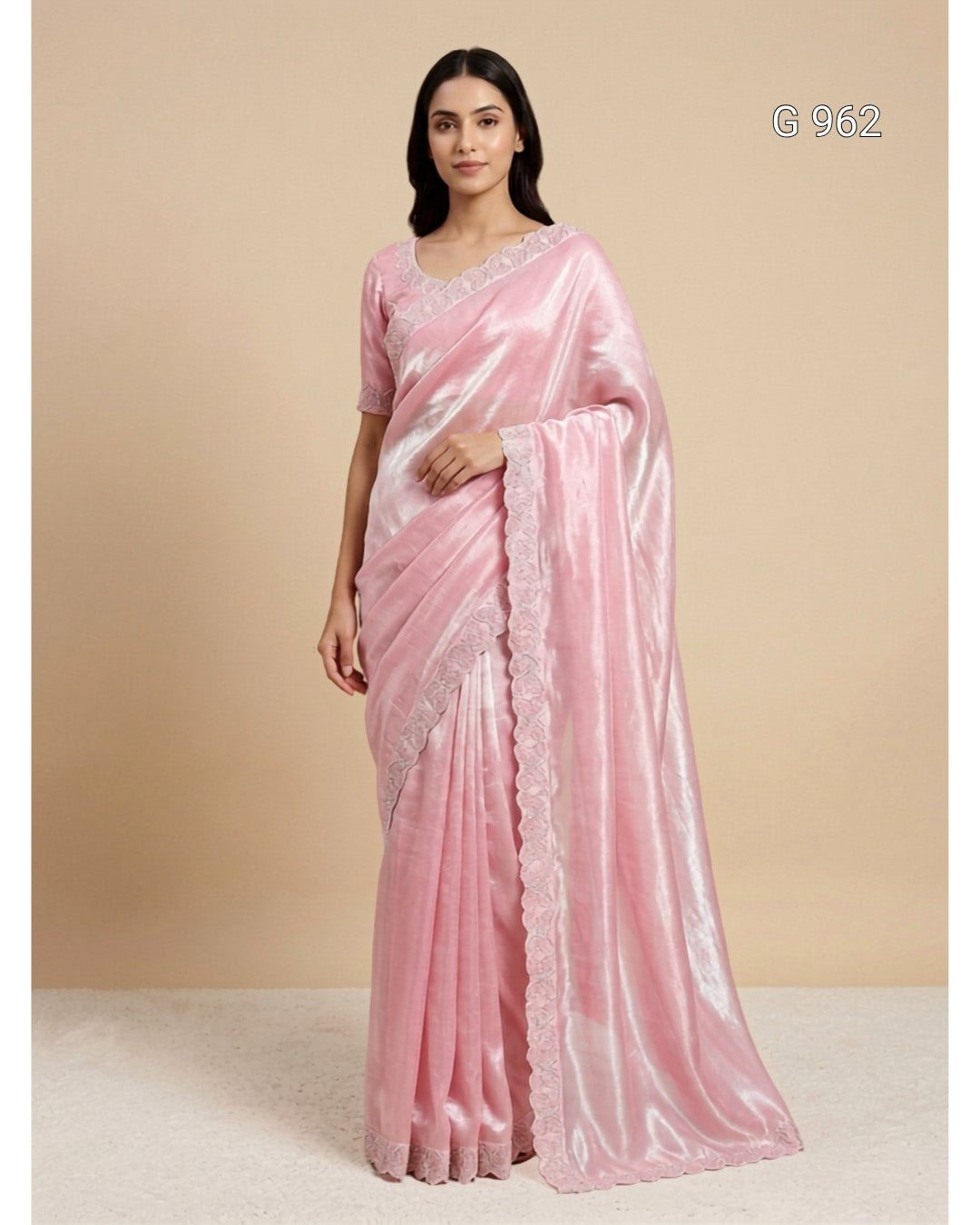 A woman modeling a light pink silk saree with a matching pink blouse, standing against a solid beige studio background. The saree has a subtle sheen and a scalloped lace border, with the code 'G 962' printed in the top right corner.