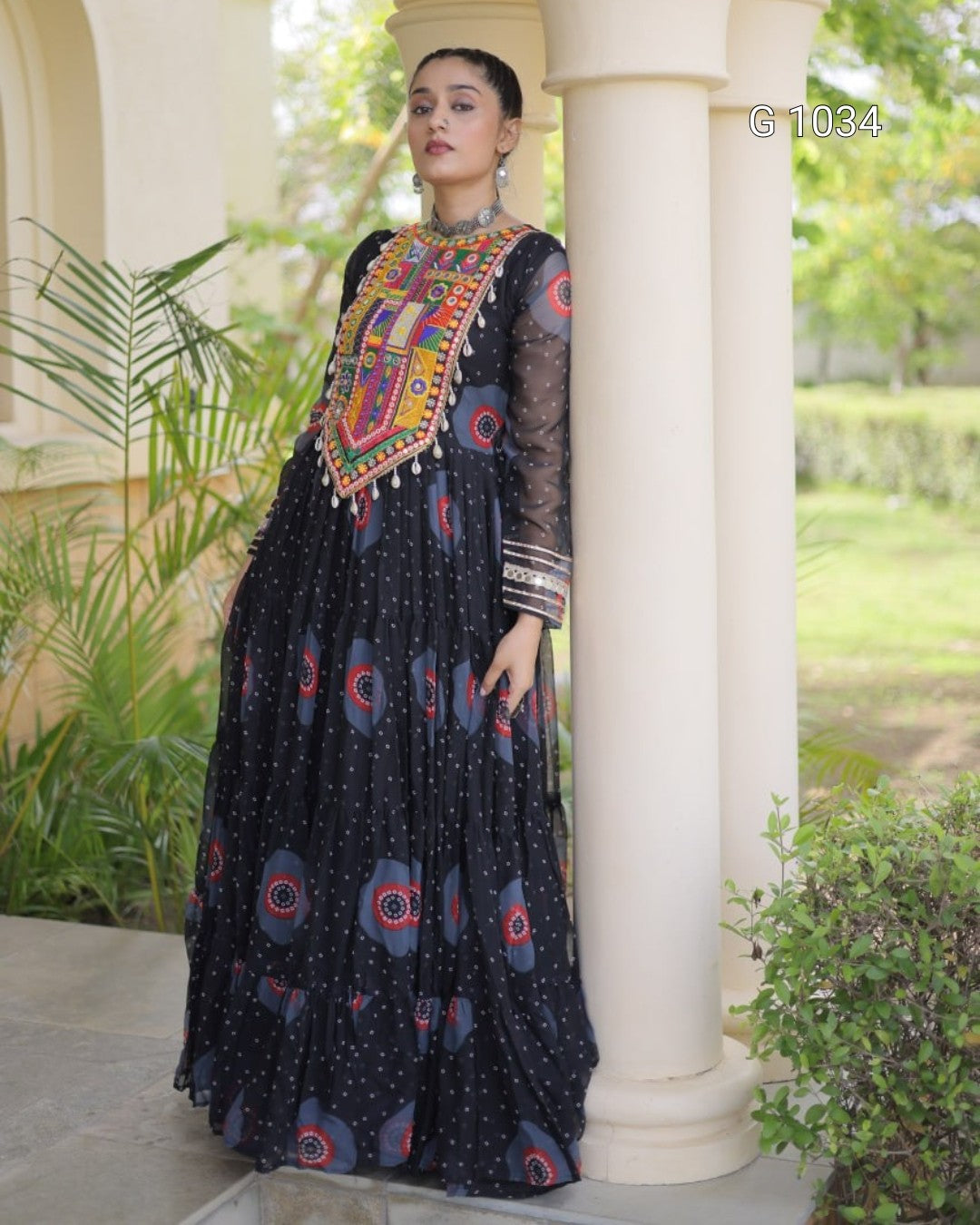 A woman models a black maxi dress with colorful geometric embroidery on the bodice and blue circular patterns on the skirt, posing outdoors next to a white column.