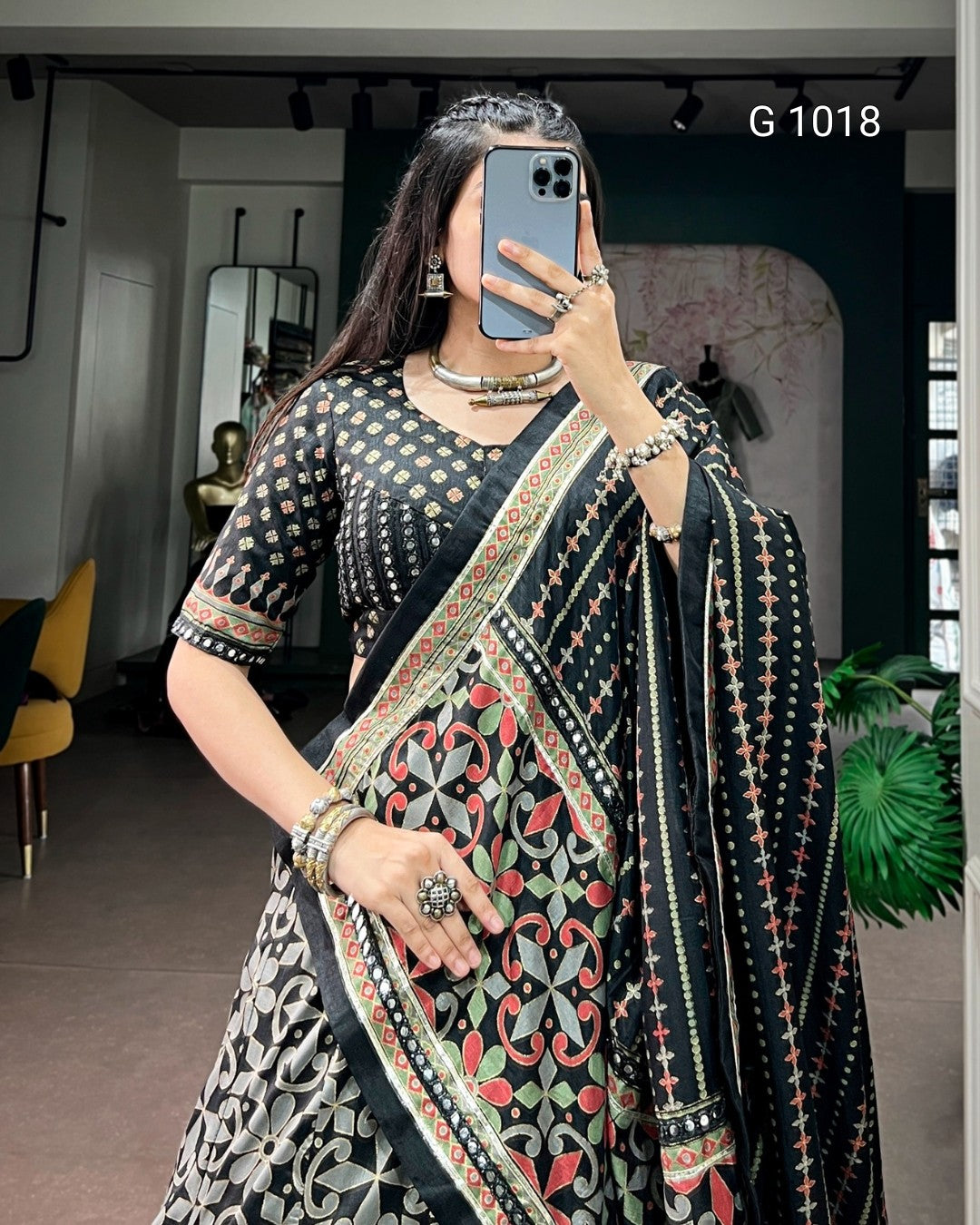 A woman takes a mirror selfie wearing a black printed saree with a heavily patterned border and matching printed blouse. The outfit features black as the dominant color with red, green, and gold accents in the floral motifs and borders.