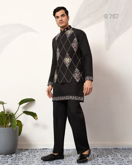 Man wearing a black embroidered traditional outfit standing in a room with a plant and patterned floor.